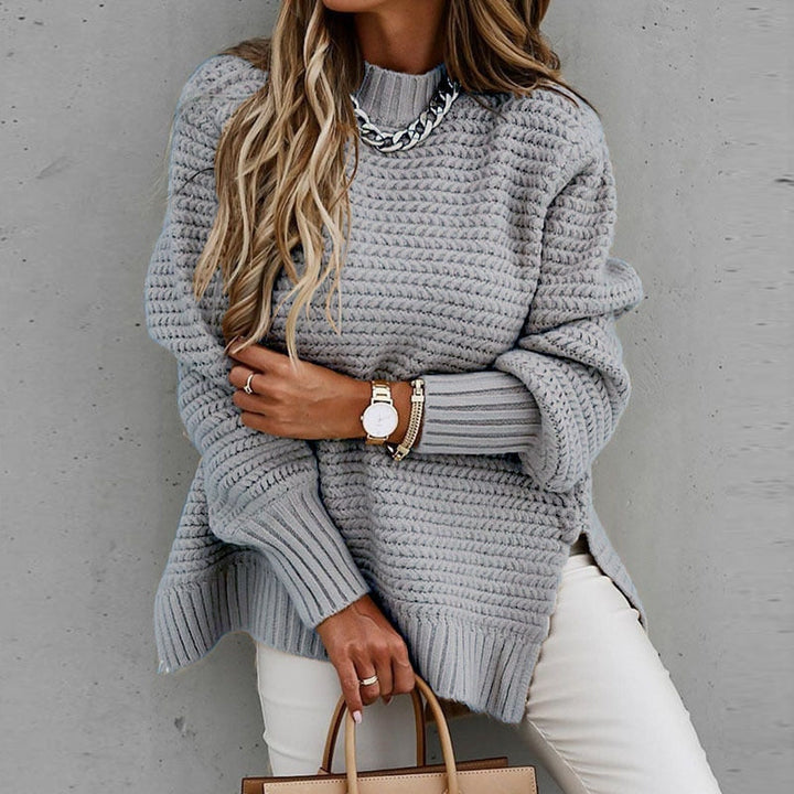 Frau in grauem Strickpullover, Kettenhalskette, weiße Hose, Armbanduhr, modisch, elegant.