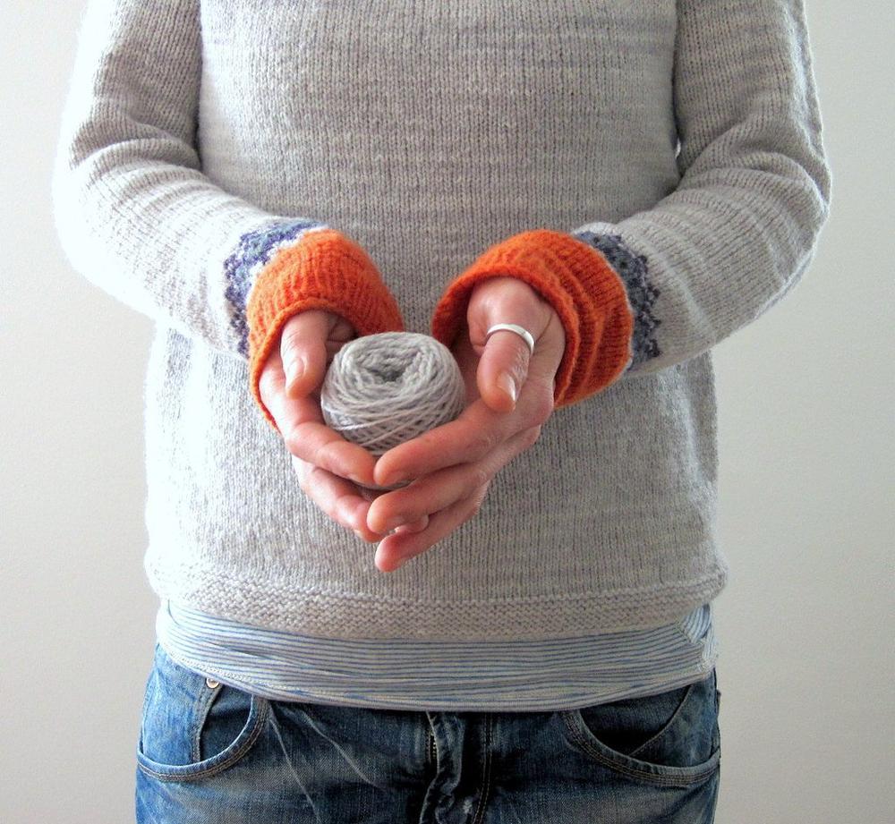 Person hält graues Wollknäuel, trägt grauen Pullover mit orangefarbenen Bündchen, blaue Jeans.