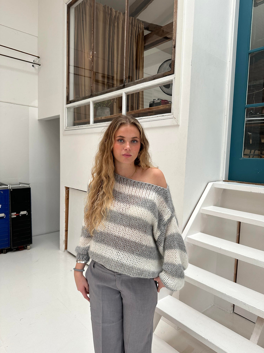 Frau in gestreiftem Pullover und grauer Hose steht vor weißer Treppe in modernem Innenraum.