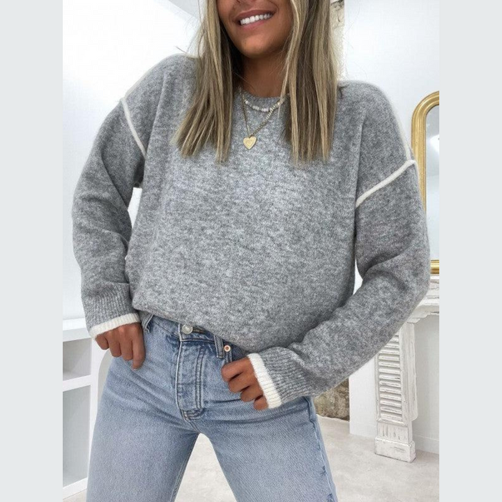 Frau trägt grauen Strickpullover, blaue Jeans, Herzanhänger; modisches Winteroutfit.