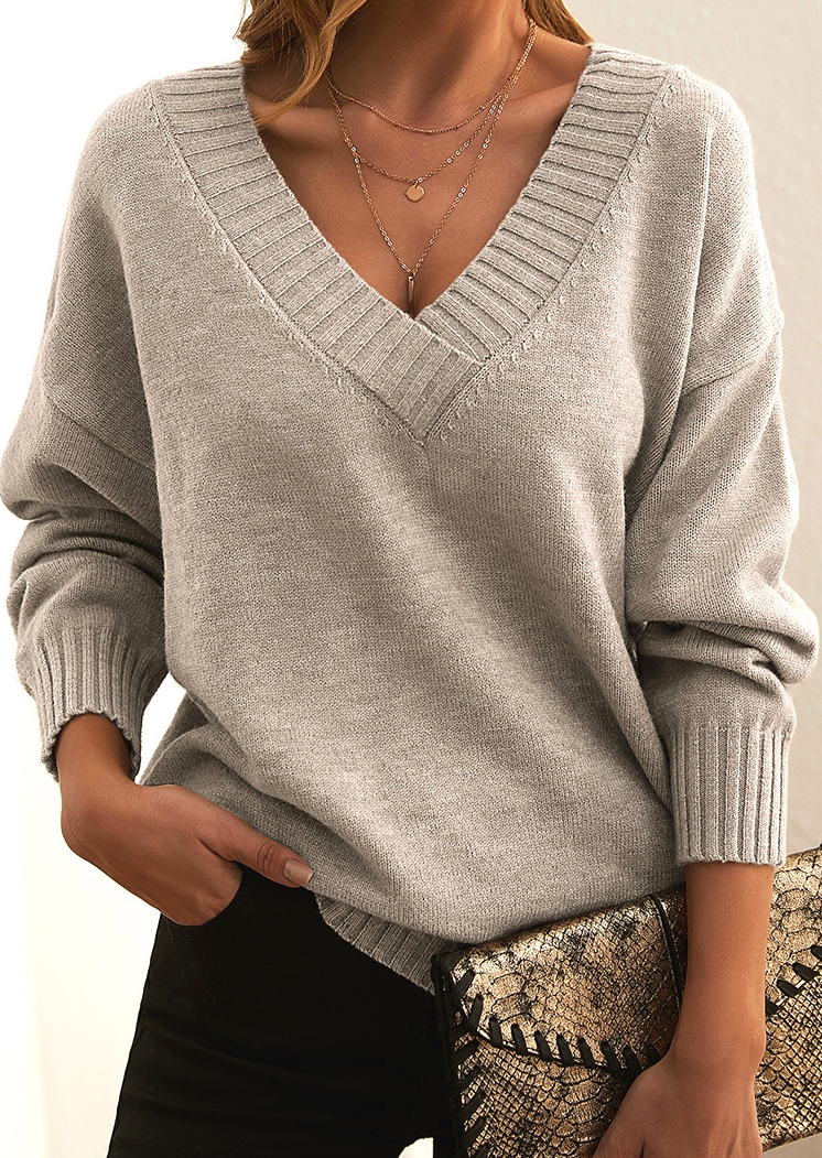 Frau in grauem V-Ausschnitt-Pullover, schwarze Hose, Schlangenhaut-Clutch, goldene Halsketten.