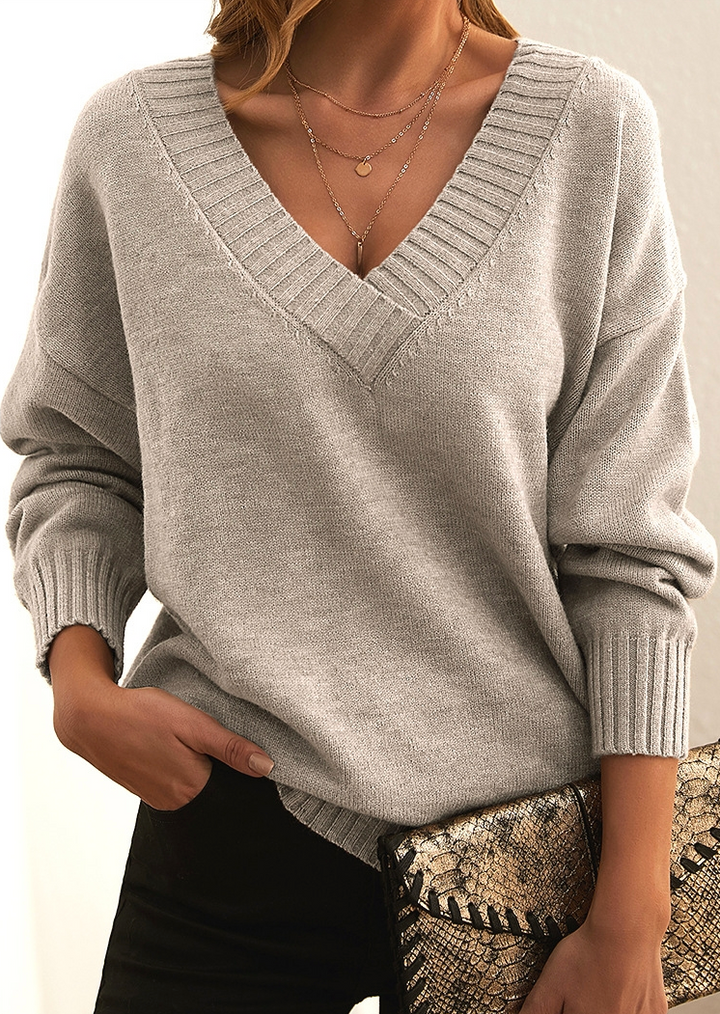 Frau in grauem V-Ausschnitt-Pullover, schwarze Hose, Schlangenhaut-Clutch, goldene Halsketten.