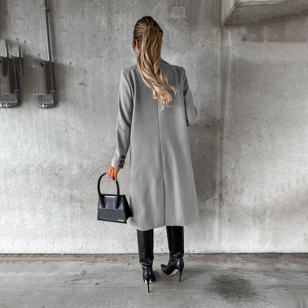 Frau in grauem Wintermantel, schwarze Stiefel, Handtasche, Betonhintergrund.