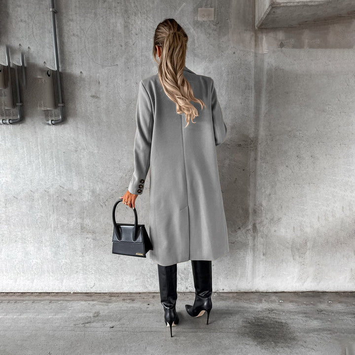 Frau in grauem Wintermantel, schwarze Stiefel, Handtasche, Betonhintergrund.