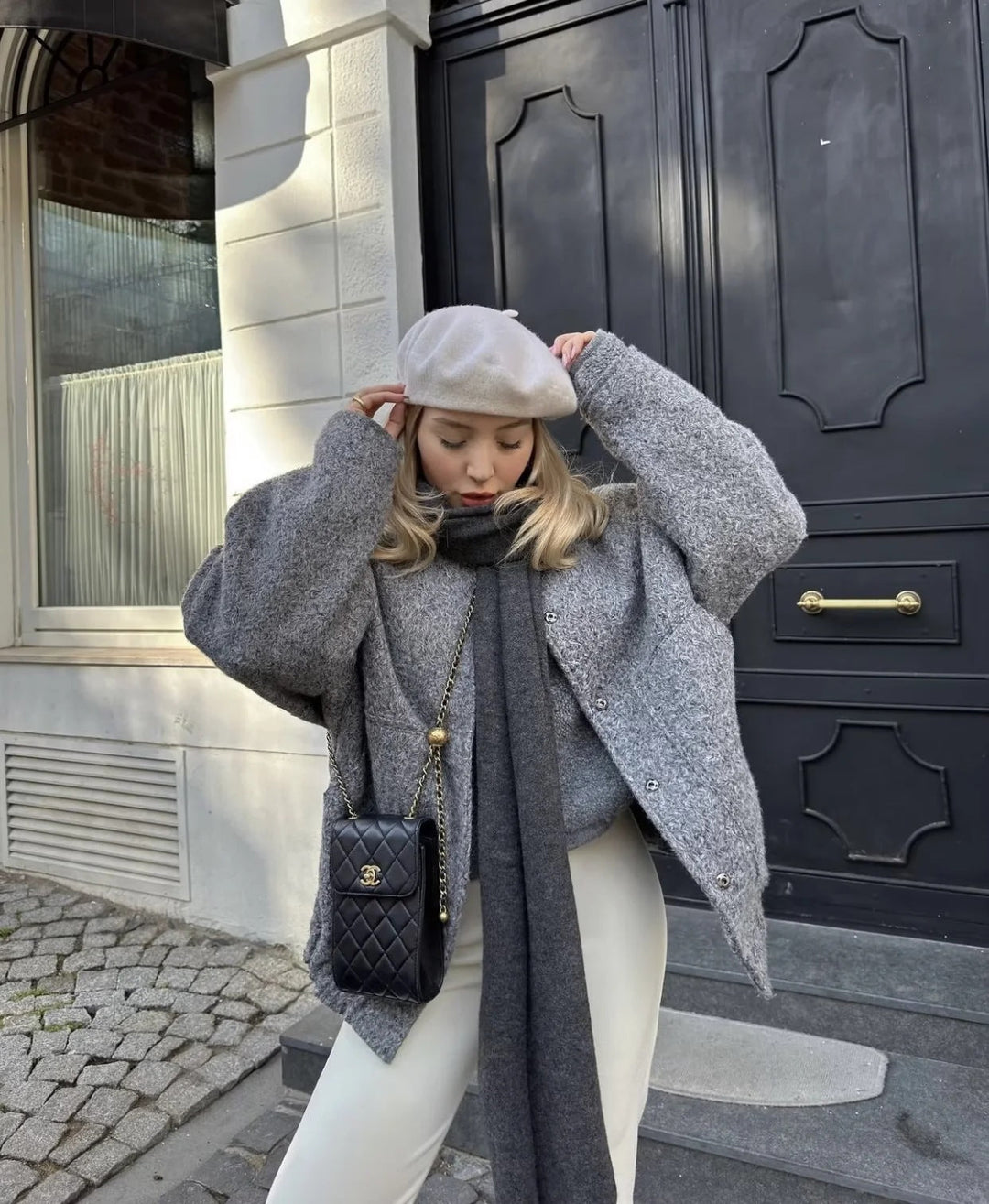 Frau in grauem Mantel und Schal, weiße Hose, schwarze Tasche, beige Baskenmütze, Stadtstraße.