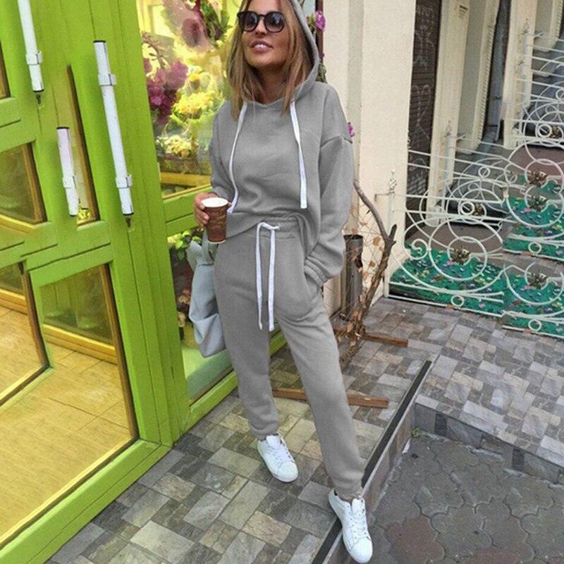 Frau in grauem Jogginganzug mit Kapuze, hält Kaffeebecher, trägt weiße Sneaker, draußen.