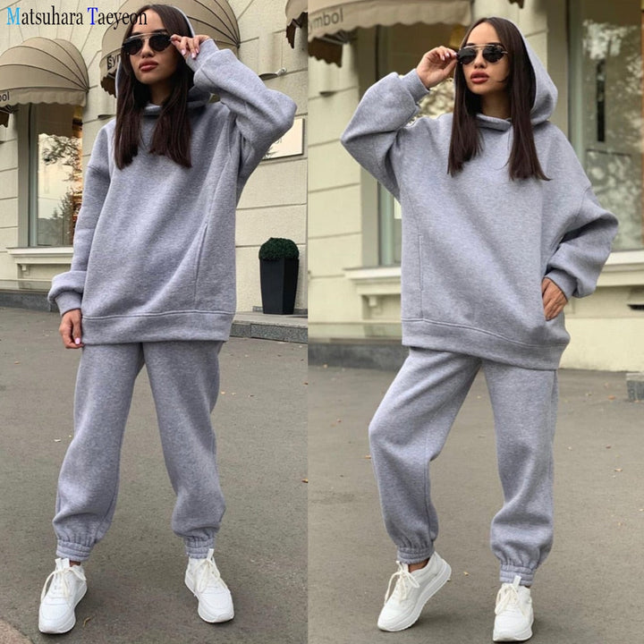 Frau in grauem Hoodie und Jogginghose, trägt Sonnenbrille, weiße Sneaker, lässiger Stil.