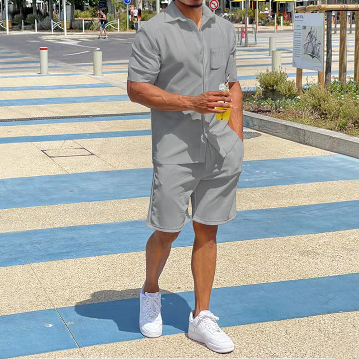 Mann in grauem Kurzarm-Overall, hält Getränk, trägt weiße Sneaker, Sommermode, urbaner Hintergrund.