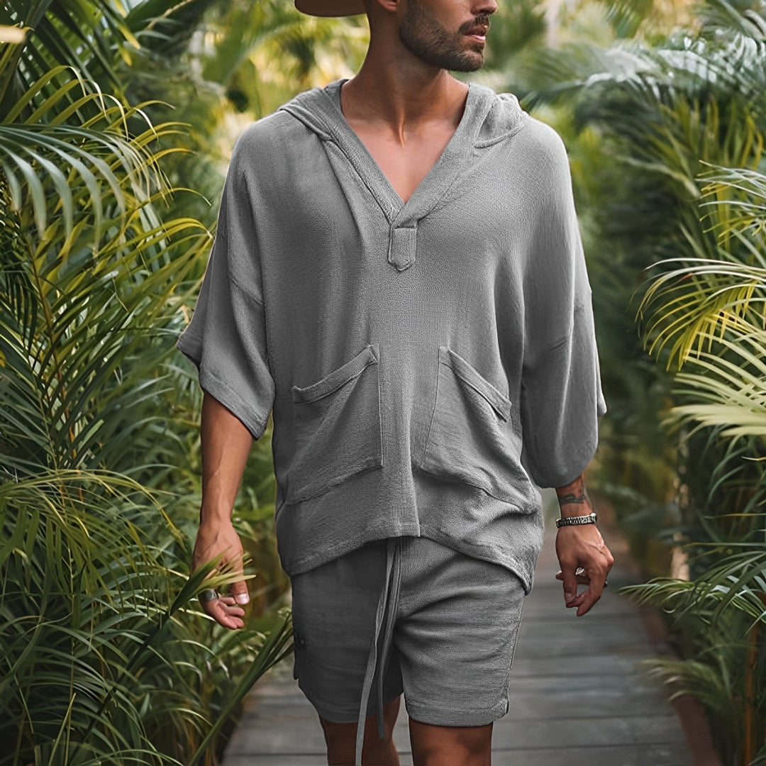 Mann in grauem Leinen-Hoodie und Shorts, lässige Sommermode, umgeben von tropischen Pflanzen.