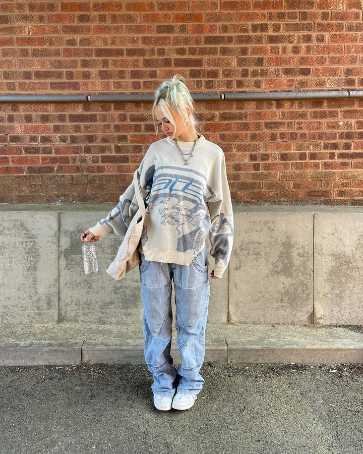 Frau in grauem Oversize-Pullover und blauer Jeans vor Backsteinmauer, lässiger Streetwear-Look.
