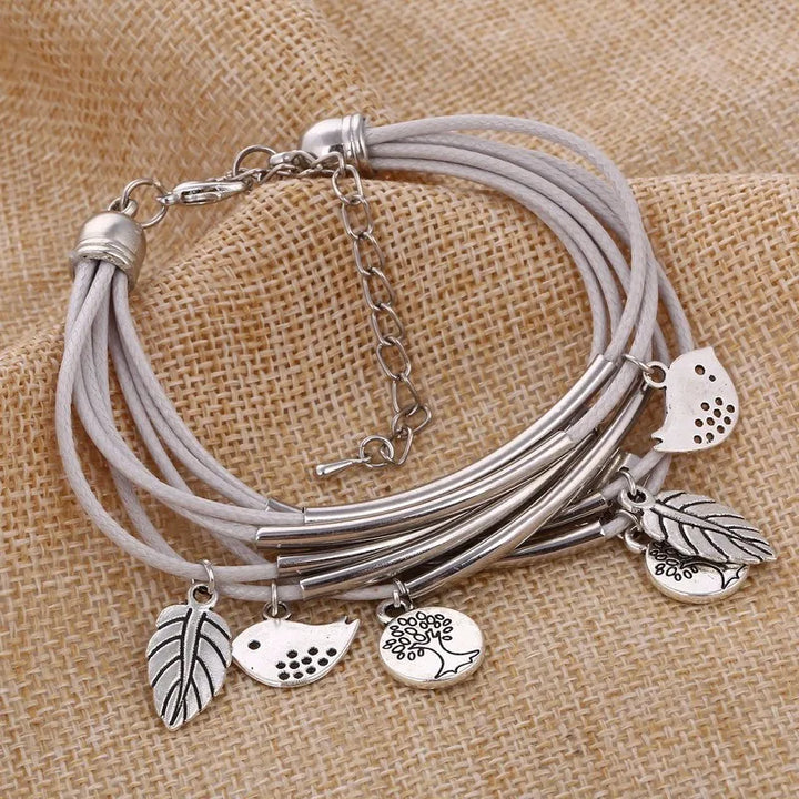 Graues Lederarmband mit silbernen Anhängern, Blätter und Vögel, auf Jutestoff.