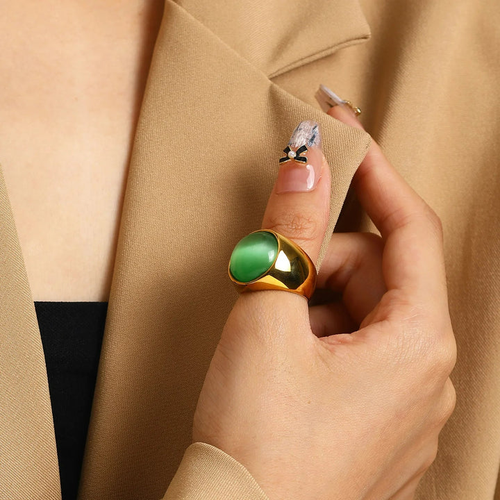 Goldener Ring mit grünem Stein, Damenhand, beiger Blazer, eleganter Schmuck, Nahaufnahme.