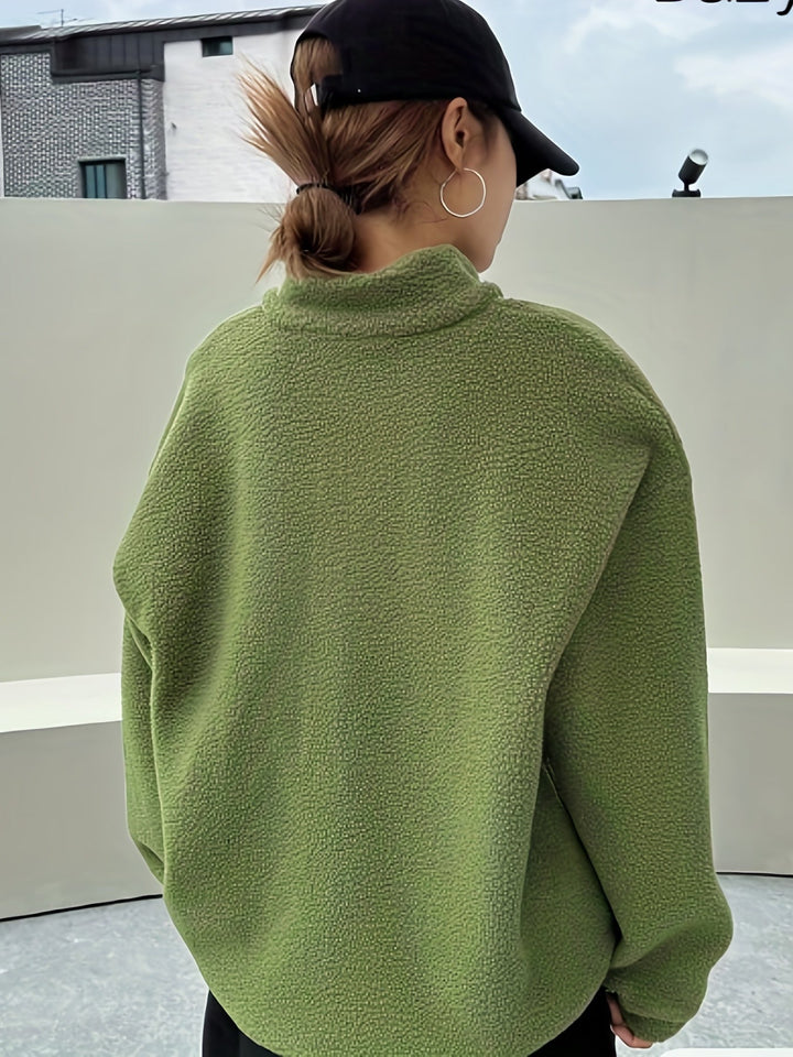 Frau in grünem Fleece-Pullover, schwarze Kappe, silberne Ohrringe, urbaner Hintergrund.