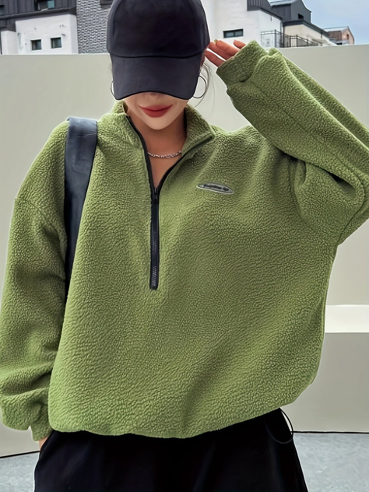 Frau in olivgrünem Fleece-Pullover mit Reißverschluss und schwarzer Kappe, urbaner Hintergrund.