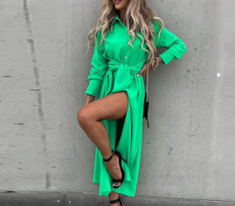 Frau in grünem Hemdkleid, knielang, mit Gürtel, schwarze Sandalen, Betonhintergrund.