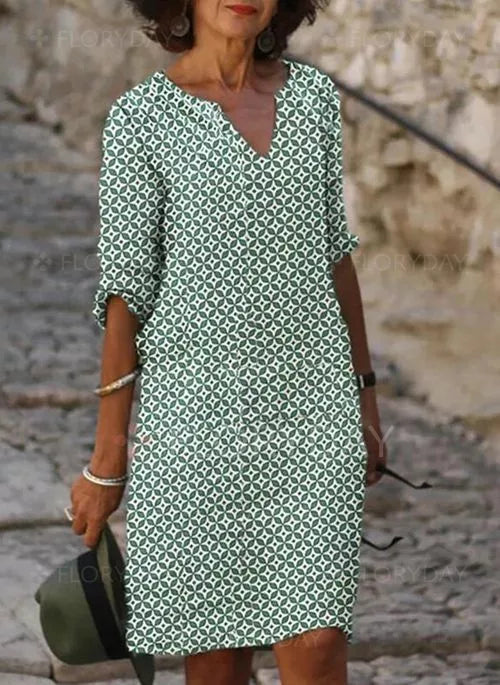 Frau in grün-weißem Sommerkleid mit geometrischem Muster, hält Hut, lässiger Stil.