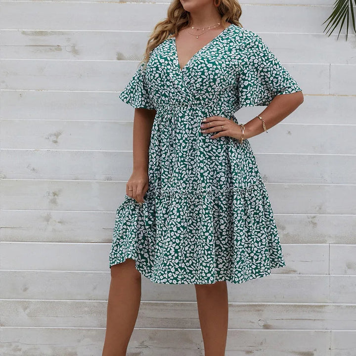 Frau in grünem Sommerkleid mit Blumenmuster vor weißer Holzwand.