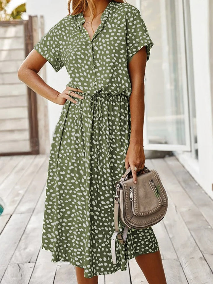 Frau in grünem Sommerkleid mit weißen Punkten, hält beige Handtasche, Holzterrasse im Hintergrund.