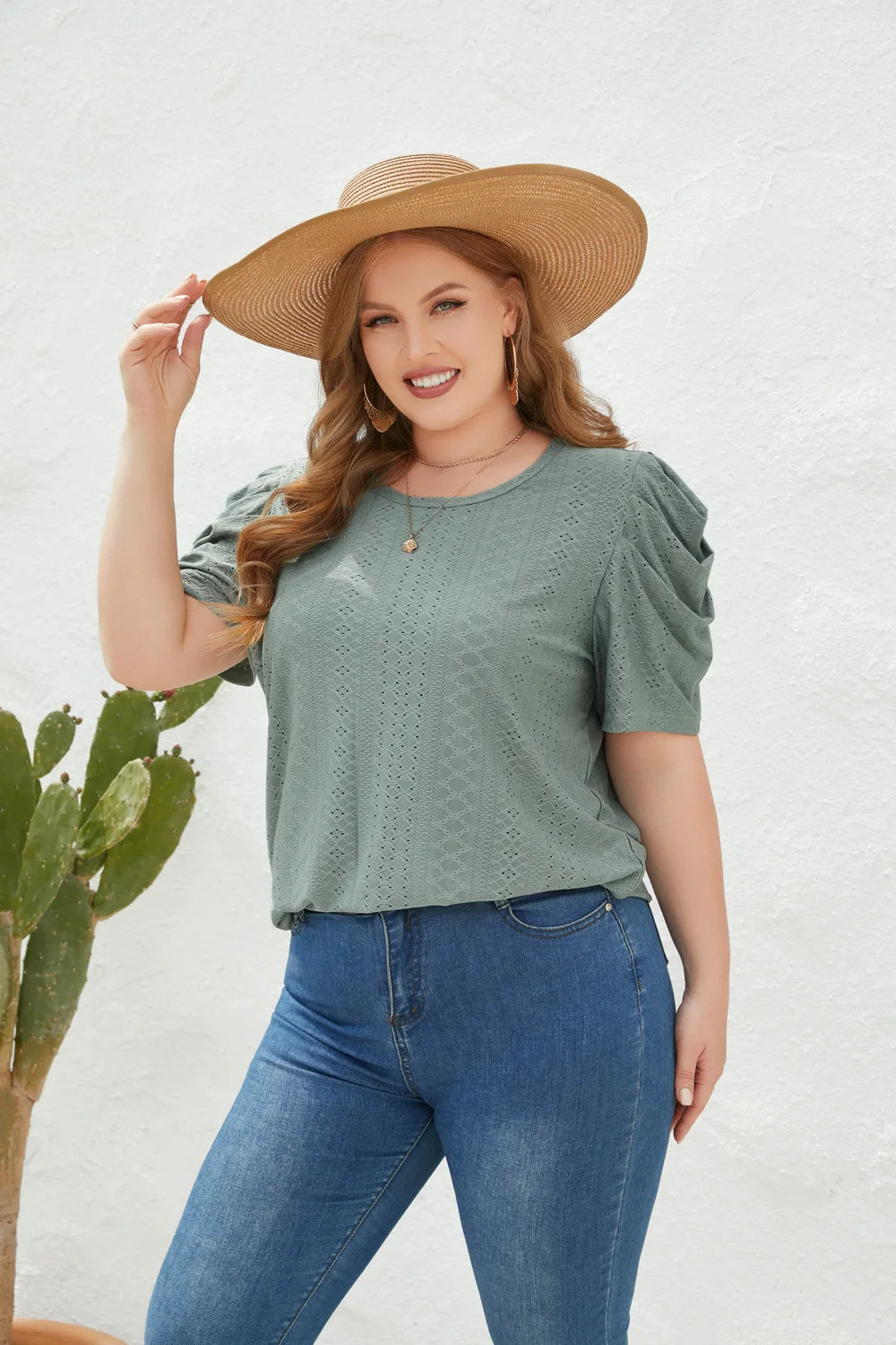 Frau in grüner Bluse und Jeans mit Strohhut, lächelnd vor weißer Wand, neben Kaktus.