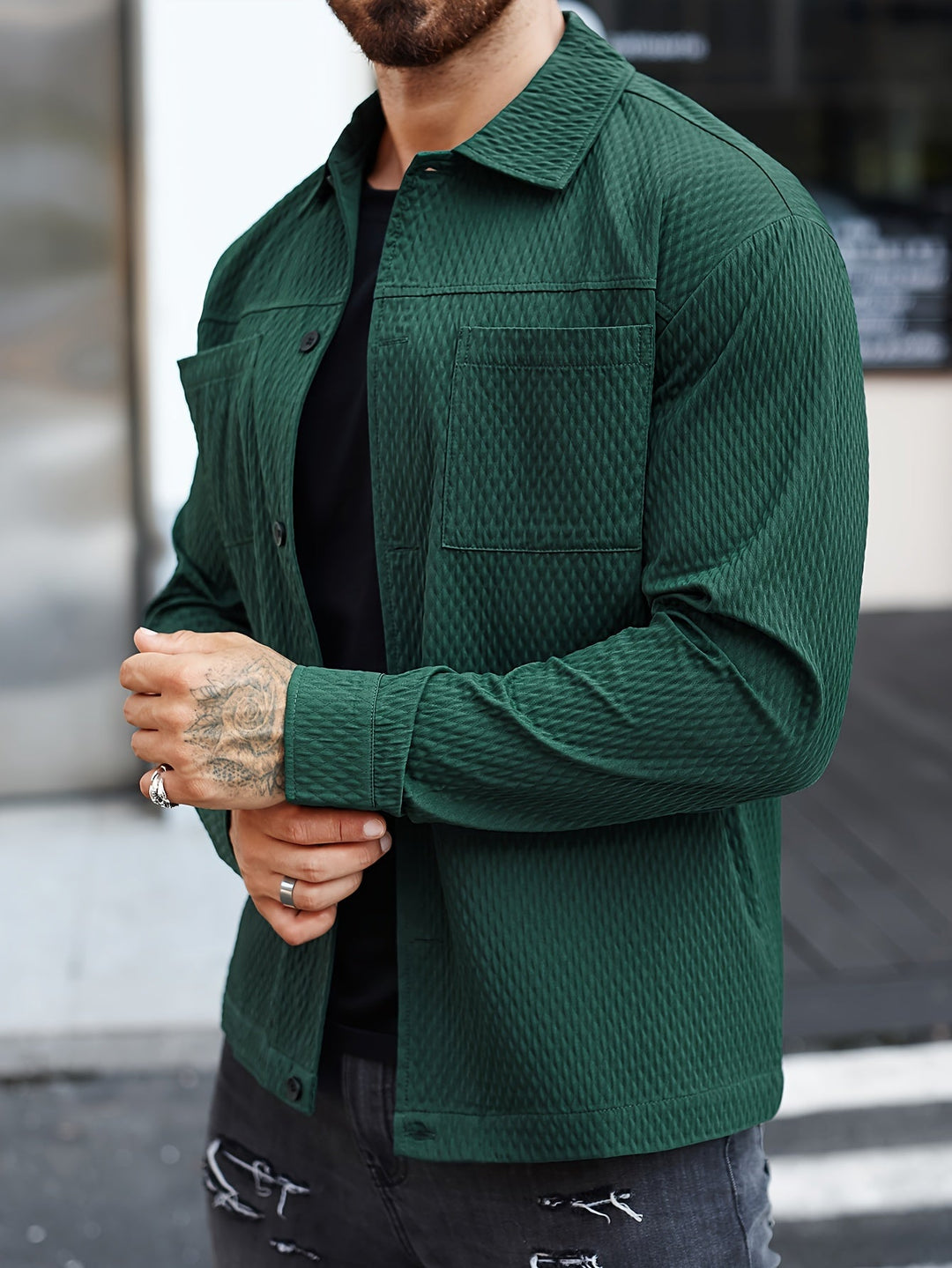 Mann in grüner Steppjacke, schwarze Jeans, tätowierte Hände, urbaner Hintergrund.