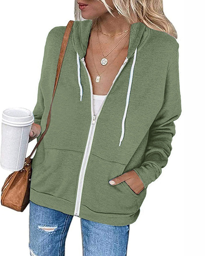 Frau in grüner Kapuzenjacke mit Reißverschluss, Kaffeebecher, Jeans, braune Tasche.
