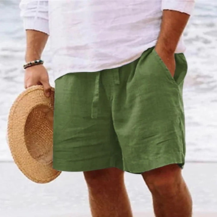 Mann in grüner Leinen-Shorts und weißem Hemd am Strand, hält Strohhut.
