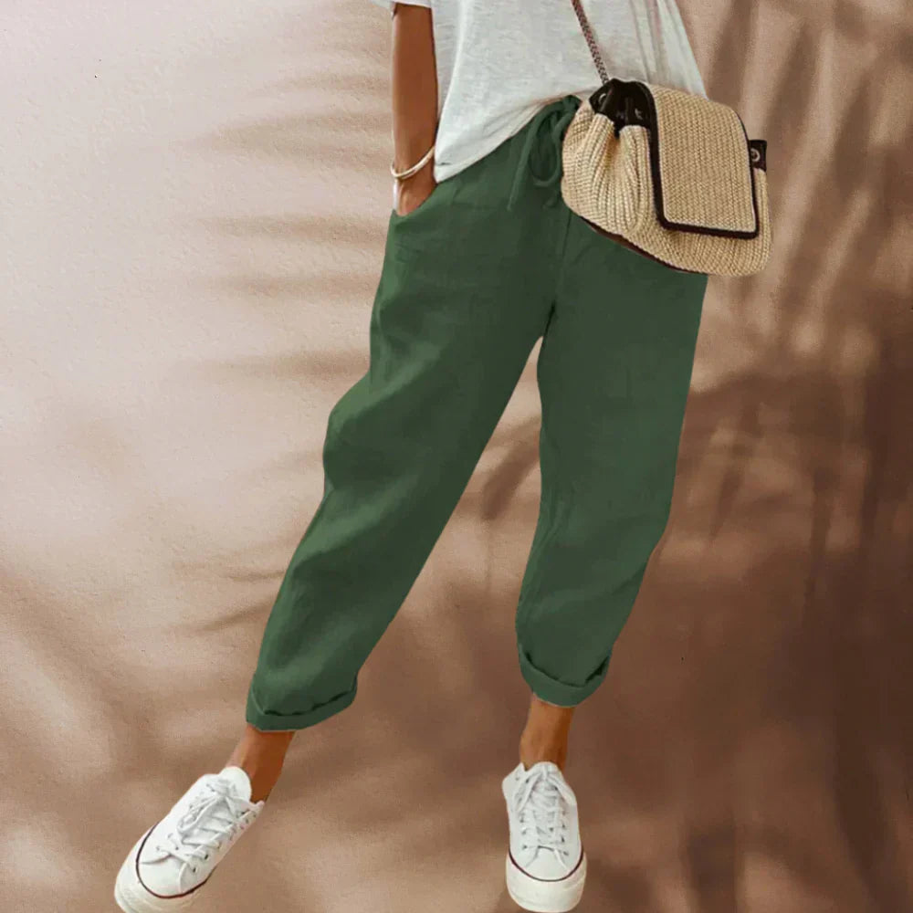 Frau in grüner Leinenhose, weißem T-Shirt, Strohtasche, weiße Sneaker, lässiger Sommerlook.