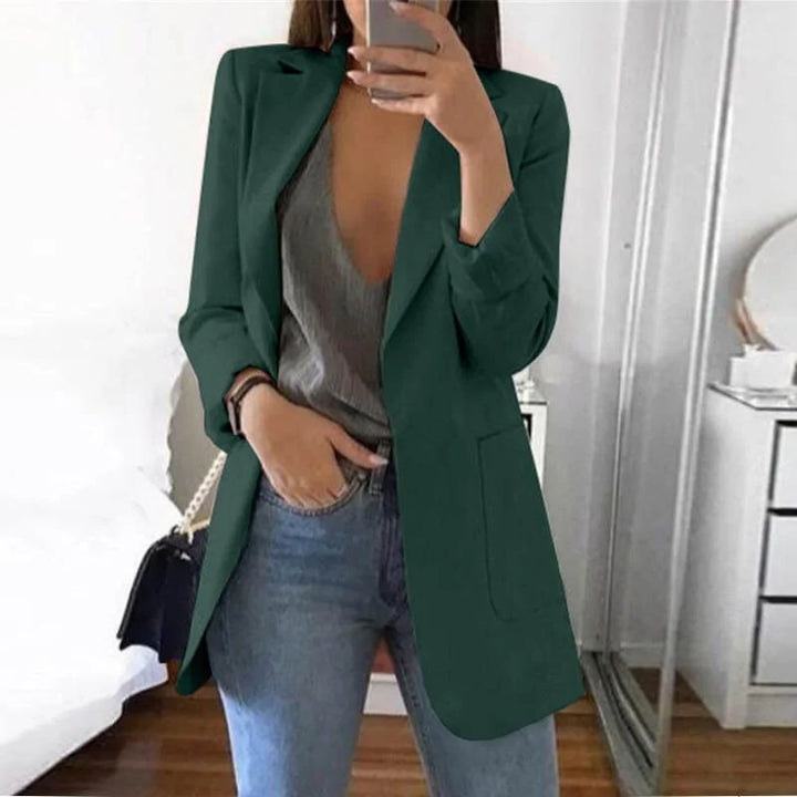 Frau in grünem Blazer, grauem Top, blauer Jeans, Spiegel-Selfie, modernes Schlafzimmer.