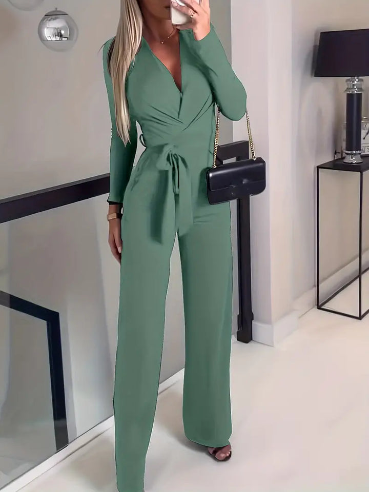 Frau in grünem Jumpsuit mit Gürtel, schwarze Umhängetasche, moderne Inneneinrichtung.