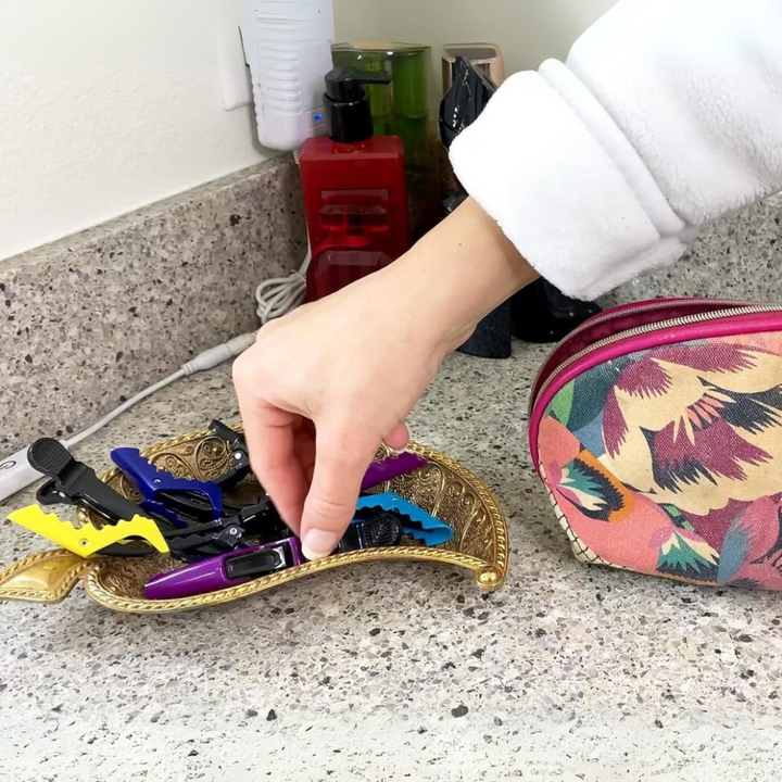 Hand greift nach bunten Haarklammern auf goldenem Tablett, bunte Kosmetiktasche daneben.