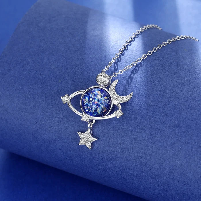 Silberne Halskette mit blauem Planeten-Anhänger, Mond und Sternen auf blauem Hintergrund.