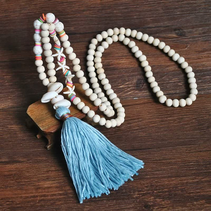 Holzperlenkette mit blauer Quaste und Muscheln auf Holztisch, handgefertigter Schmuck.