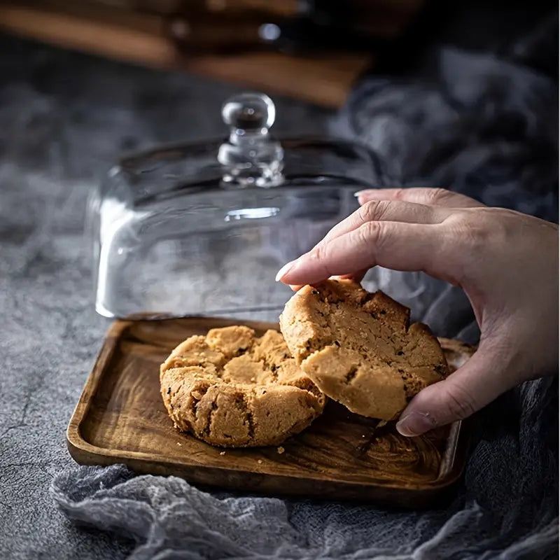 Hand greift nach Erdnussbutterkeks auf Holztablett unter Glasglocke, rustikaler Hintergrund.