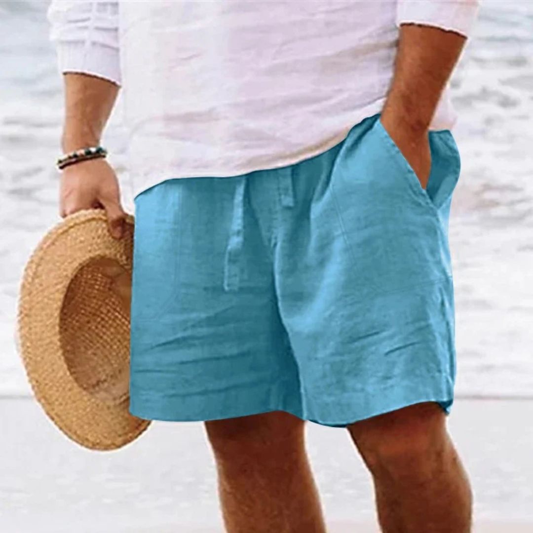 Mann in blauer Leinen-Shorts und weißem Hemd am Strand, hält Strohhut.