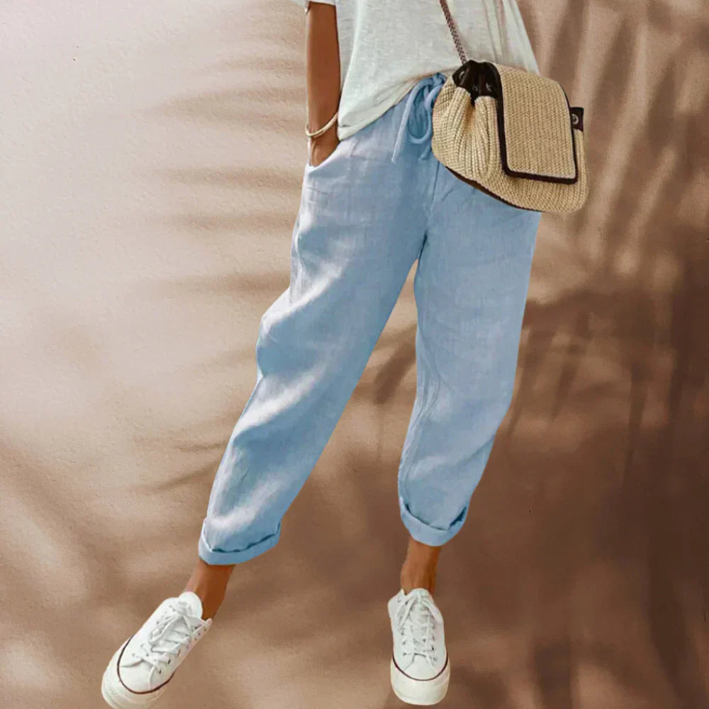 Frau in hellblauer Leinenhose, weißem T-Shirt, weißen Sneakers, Strohtasche, lässiger Look.
