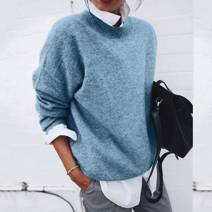 Frau in blauem Strickpullover, weißer Bluse, grauer Hose, schwarze Tasche, lässiger Stil.