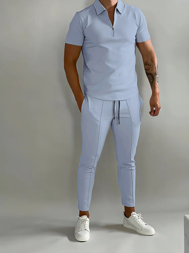 Mann in hellblauem Trainingsanzug und weißen Sneakers vor grauem Hintergrund.
