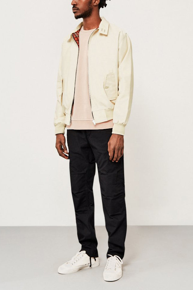 Mann in cremefarbener Jacke, schwarzer Hose und weißen Sneakers vor neutralem Hintergrund.