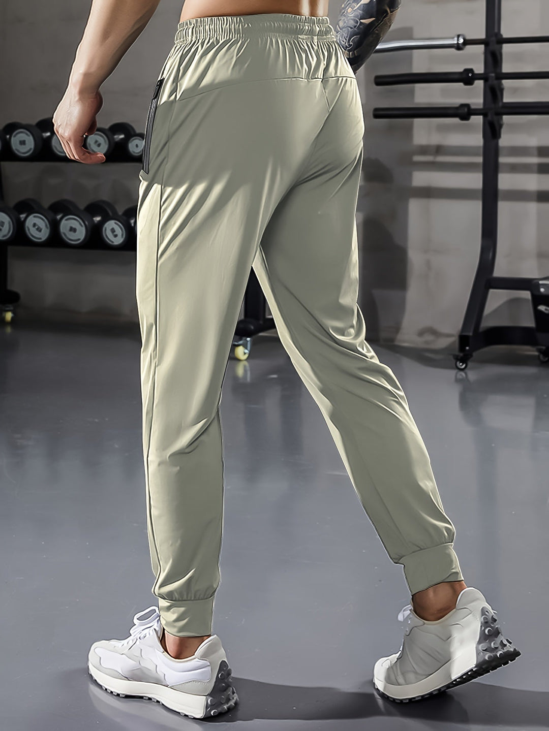 Mann in hellgrauer Jogginghose im Fitnessstudio, Hanteln im Hintergrund.