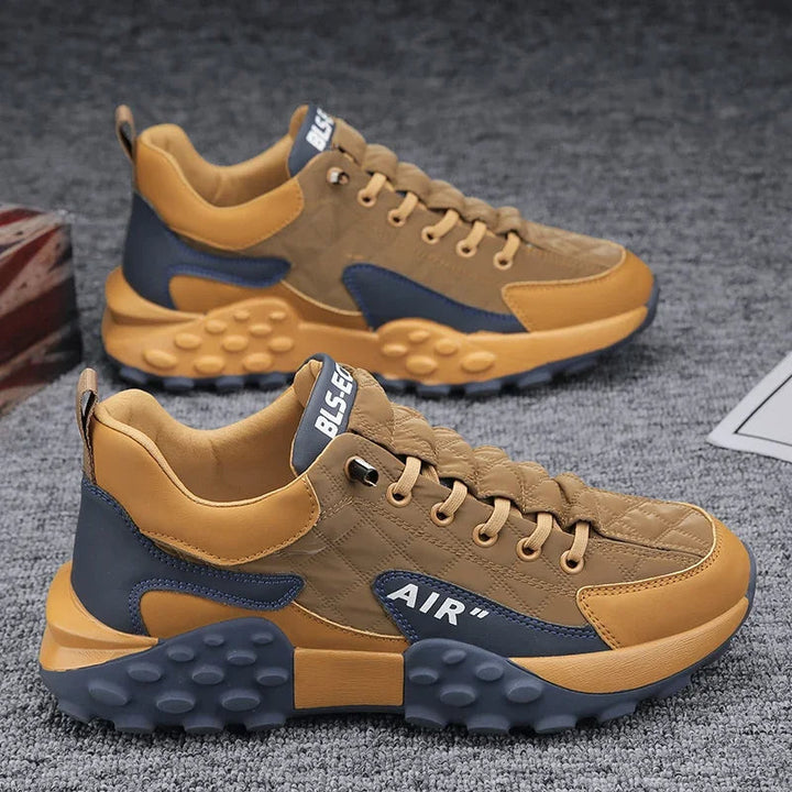 Braune und graue Herren-Sportschuhe, atmungsaktiv, mit Luftkissen-Sohle auf grauem Teppich.