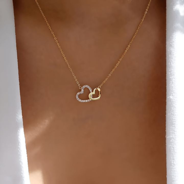 Goldene Halskette mit zwei Herzanhängern, einer mit Diamanten, auf Hauthintergrund.