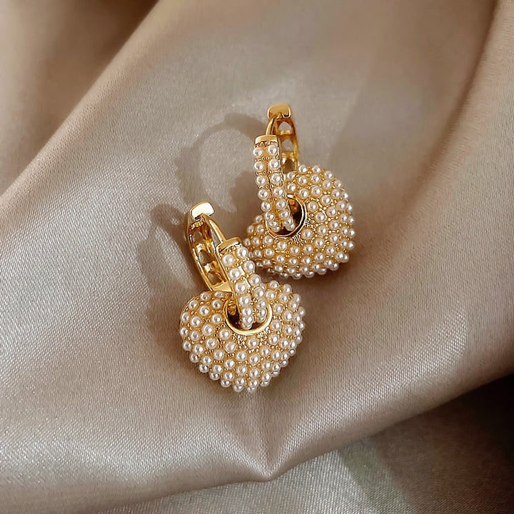 Goldene Herz-Ohrringe mit Perlen auf beigem Stoff, eleganter Schmuck, modisches Accessoire.