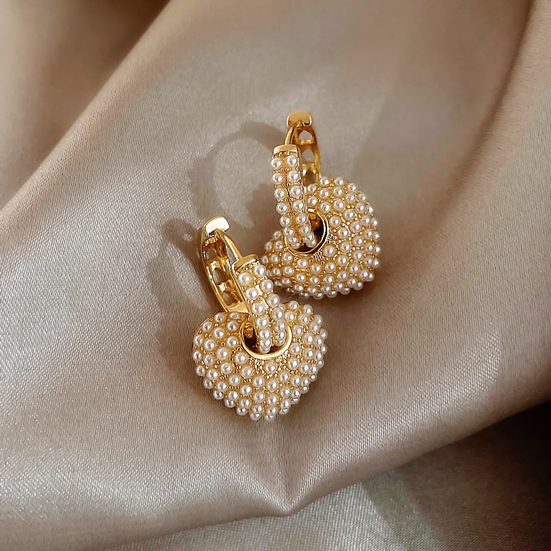 Goldene Herz-Ohrringe mit Perlen auf beigem Stoff, eleganter Schmuck, modische Accessoires.