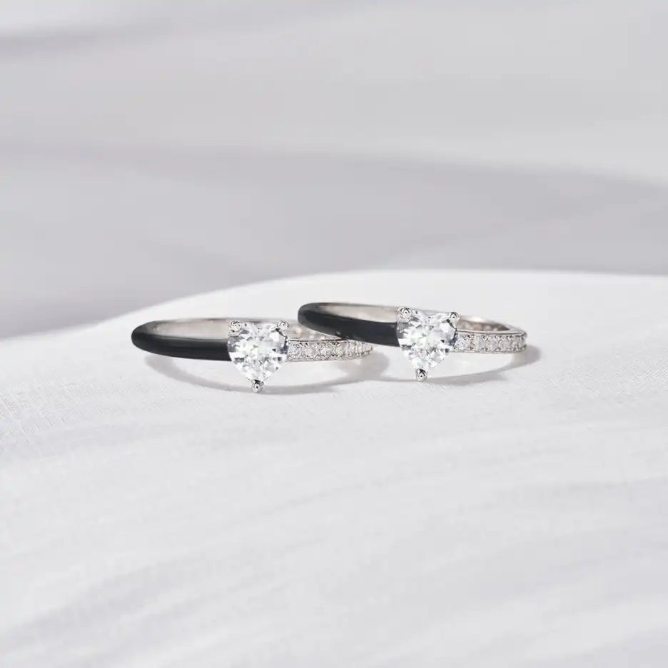 Zwei silberne Verlobungsringe mit Herz-Diamanten und schwarzen Details auf weißem Hintergrund.