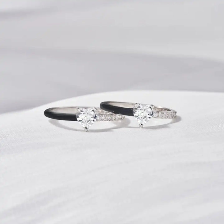 Zwei silberne Verlobungsringe mit Herz-Diamanten und schwarzen Details auf weißem Hintergrund.