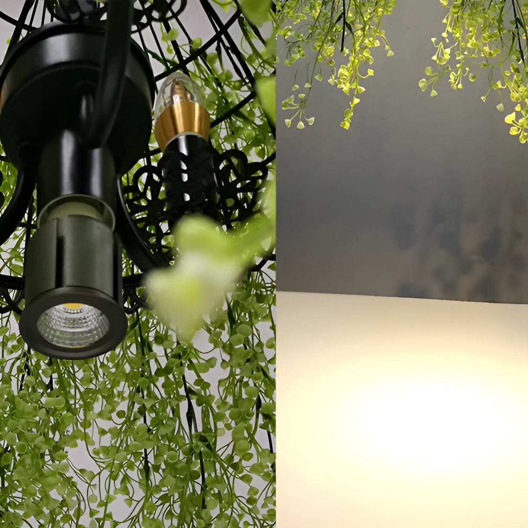 Detail černé kovové objímky s žárovkami a LED reflektorkem mezi zelenými lístky, vedle zářící plochy 9