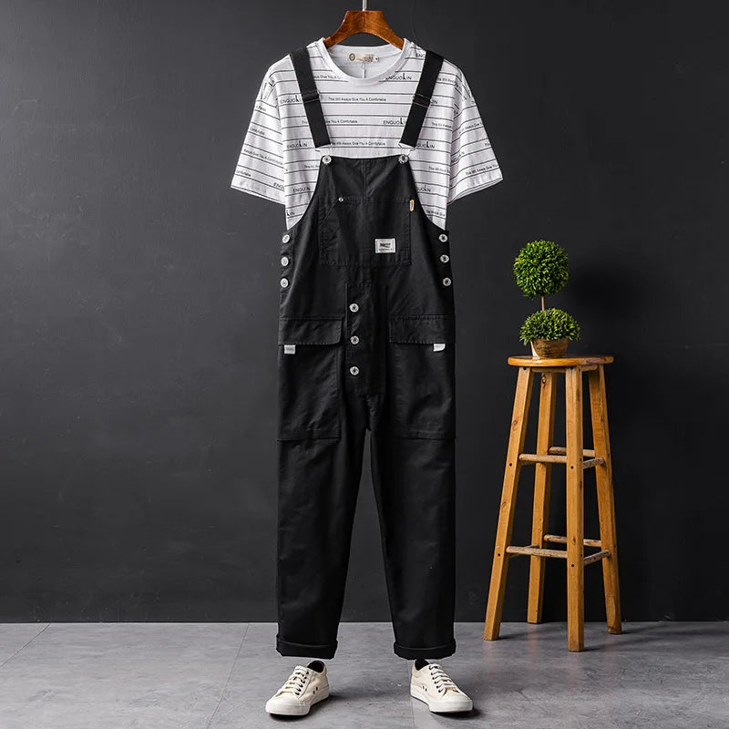 Schwarze Herren-Latzhose mit Taschen, lässig, Sommermode, auf Kleiderbügel vor dunklem Hintergrund.