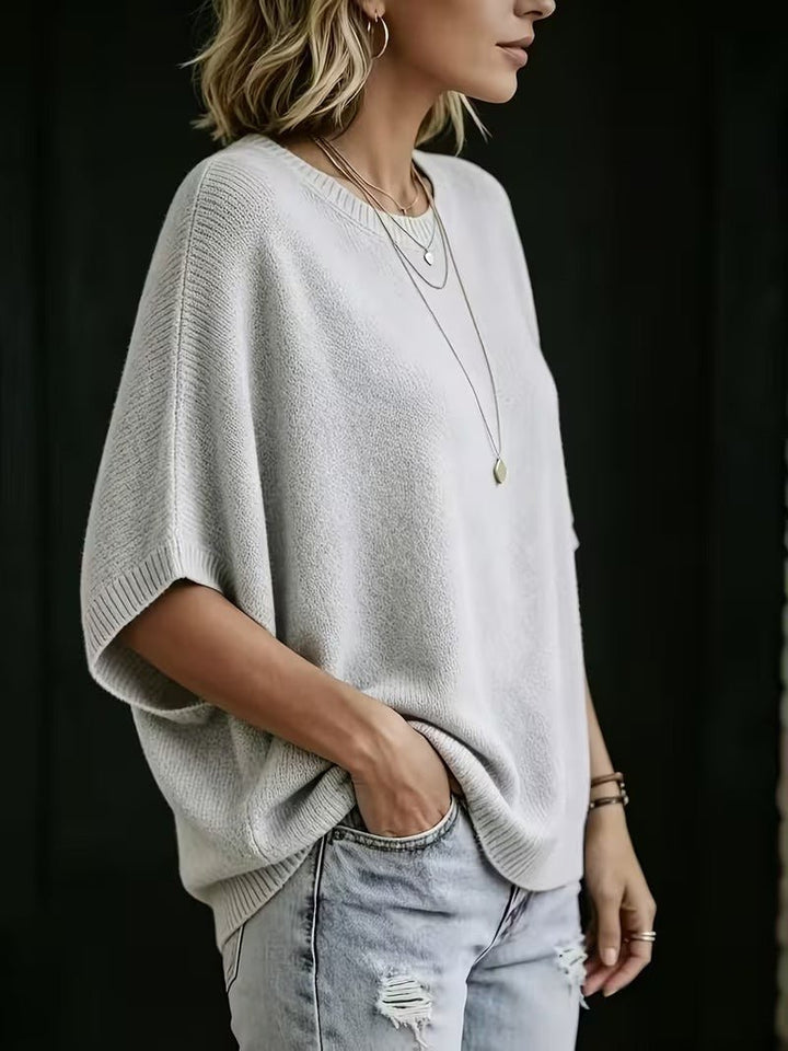 Šedý pletený top s krátkými netopýřími rukávy na modelce z boku, ruka v kapse džínů. 2