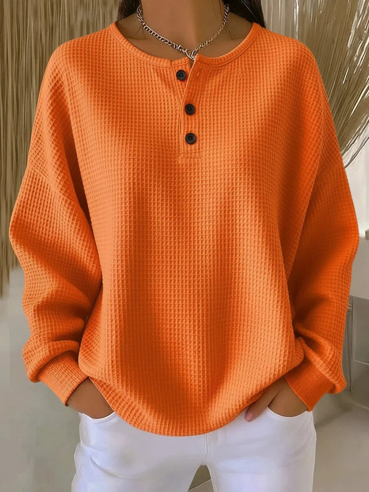 Jednobarevný oranžový vaflový svetr se třemi černými knoflíky pod kulatým výstřihem, nošený s bílými kalhotami. 7