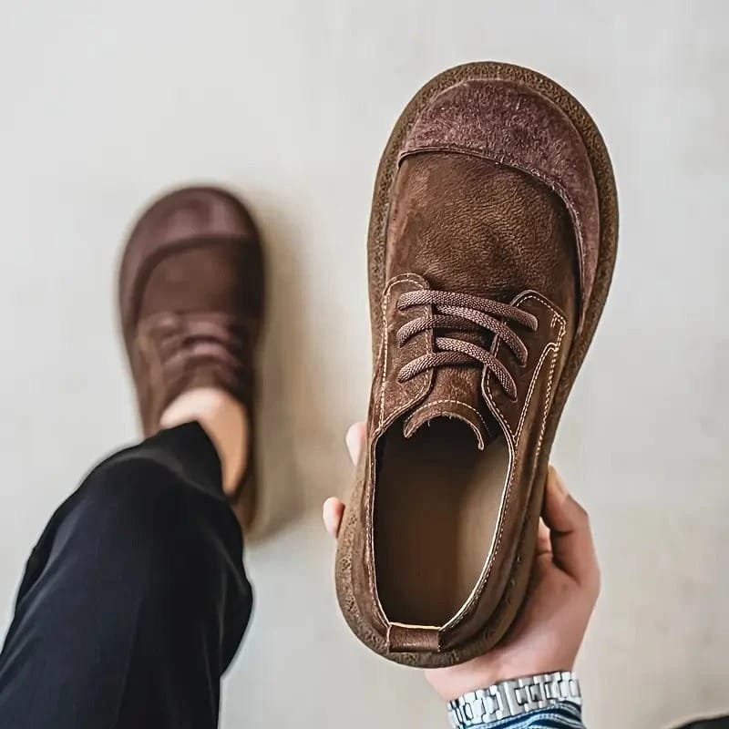 Tmavě hnědá šněrovací bota z pohledu shora v ruce, druhá bota na noze v černých kalhotách. 2